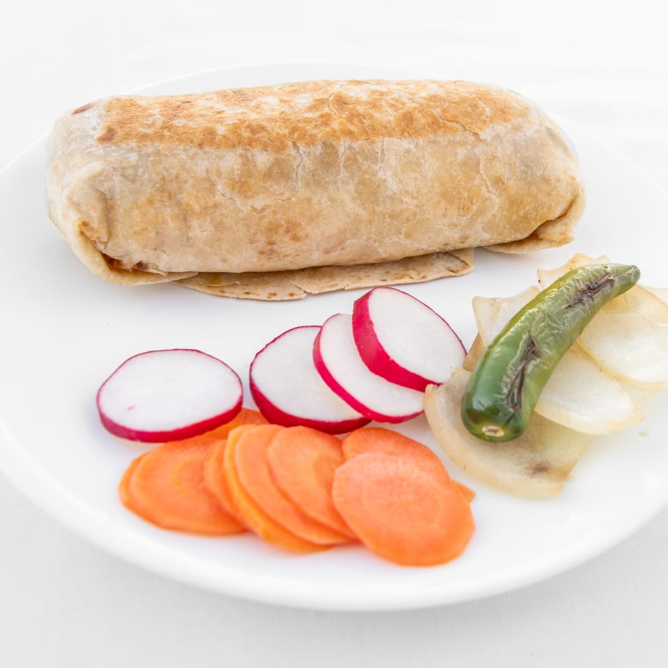 A large, golden-brown burrito on a white plate with sliced radishes, carrots, and grilled onions.