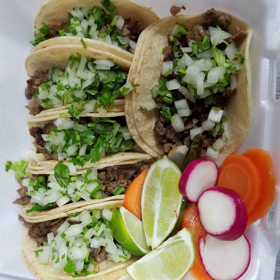 Six carne asada tacos with cilantro and onions, served with lime wedges, radishes, and carrots.