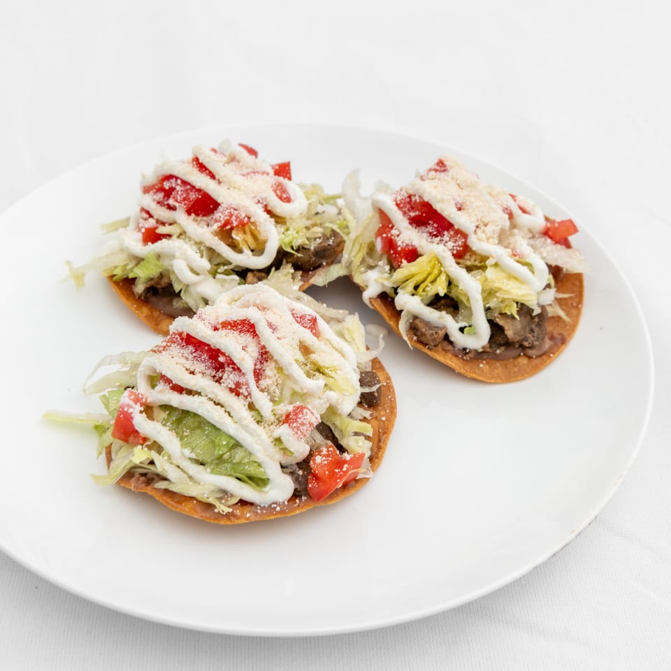 Three Mexican tostadas with meat, lettuce, tomatoes, crema, and cheese on a white plate.