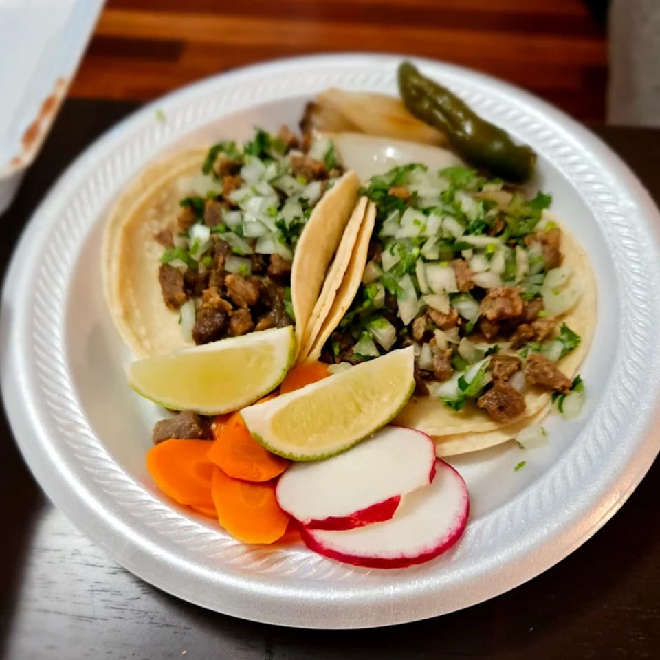 Three Mexican street tacos with meat, onions, cilantro, lime, carrots, radishes, and a grilled pepper.